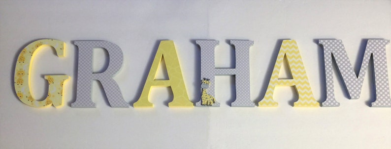 May include: Wooden letters spelling the name "GRAHAM" in yellow and grey with a giraffe pattern on the "G" and "H".