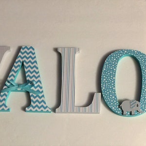 May include: Wooden letters spelling the name "AVALON" in blue, gray, and white. Each letter has a different pattern, including elephants, chevron, and polka dots. The letter "O" has a small elephant figurine attached.