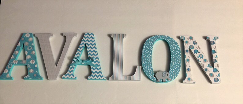 May include: Wooden letters spelling the name "AVALON" in blue, gray, and white. Each letter has a different pattern, including elephants, chevron, and polka dots. The letter "O" has a small elephant figurine attached.