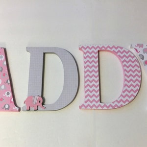 May include: Wooden letters spelling "ADDY" in pink and gray. The letters are decorated with different patterns, including pink and gray elephants, gray and white checkered, and pink and white chevron.