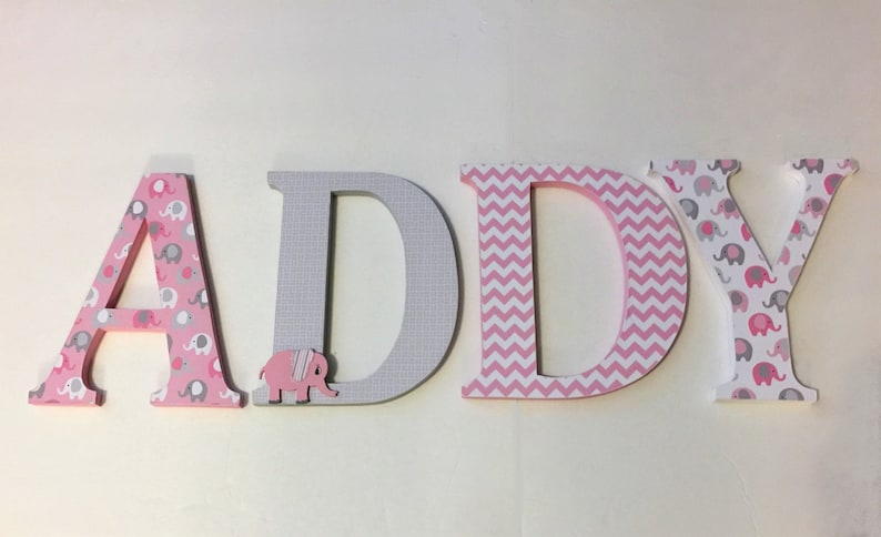 May include: Wooden letters spelling "ADDY" in pink and gray. The letters are decorated with different patterns, including pink and gray elephants, gray and white checkered, and pink and white chevron.