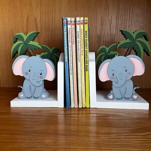 Peut inclure: Des serre-livres en forme d'&eacute;l&eacute;phant blancs avec des &eacute;l&eacute;phants gris et des c&oelig;urs roses. Les serre-livres tiennent une rang&eacute;e de livres pour enfants avec des titres comme "Cendrillon", "Pinocchio" et "Brer Rabbit Saves His Skin".