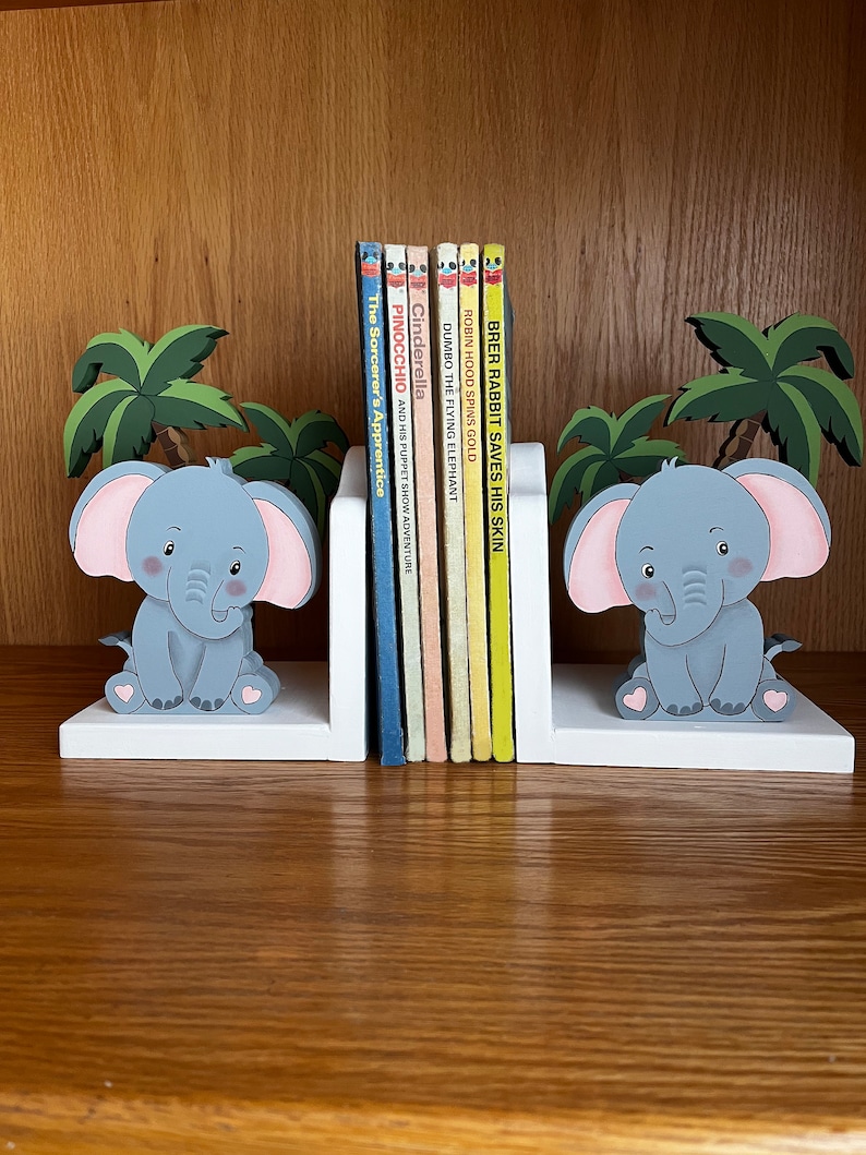Peut inclure: Des serre-livres en forme d'&eacute;l&eacute;phant blancs avec des &eacute;l&eacute;phants gris et des c&oelig;urs roses. Les serre-livres tiennent une rang&eacute;e de livres pour enfants avec des titres comme "Cendrillon", "Pinocchio" et "Brer Rabbit Saves His Skin".