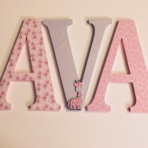 May include: Wooden letters spelling "AVA" in pink and grey with a small giraffe figurine in the middle. The letters are decorated with a pink and white giraffe print.