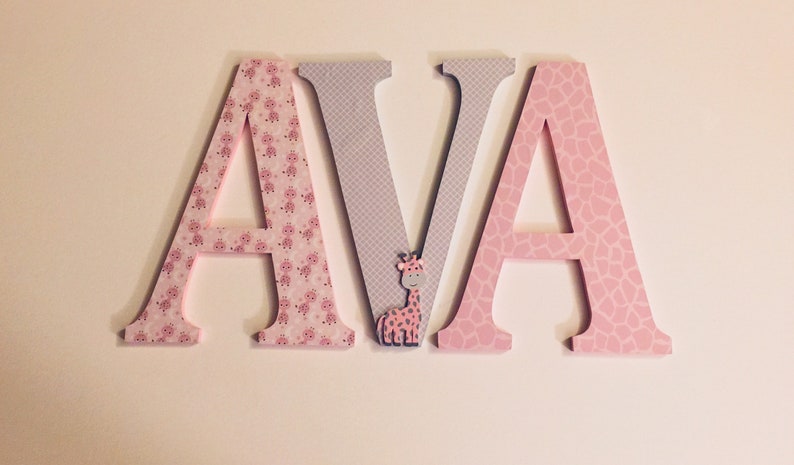 May include: Wooden letters spelling "AVA" in pink and grey with a small giraffe figurine in the middle. The letters are decorated with a pink and white giraffe print.