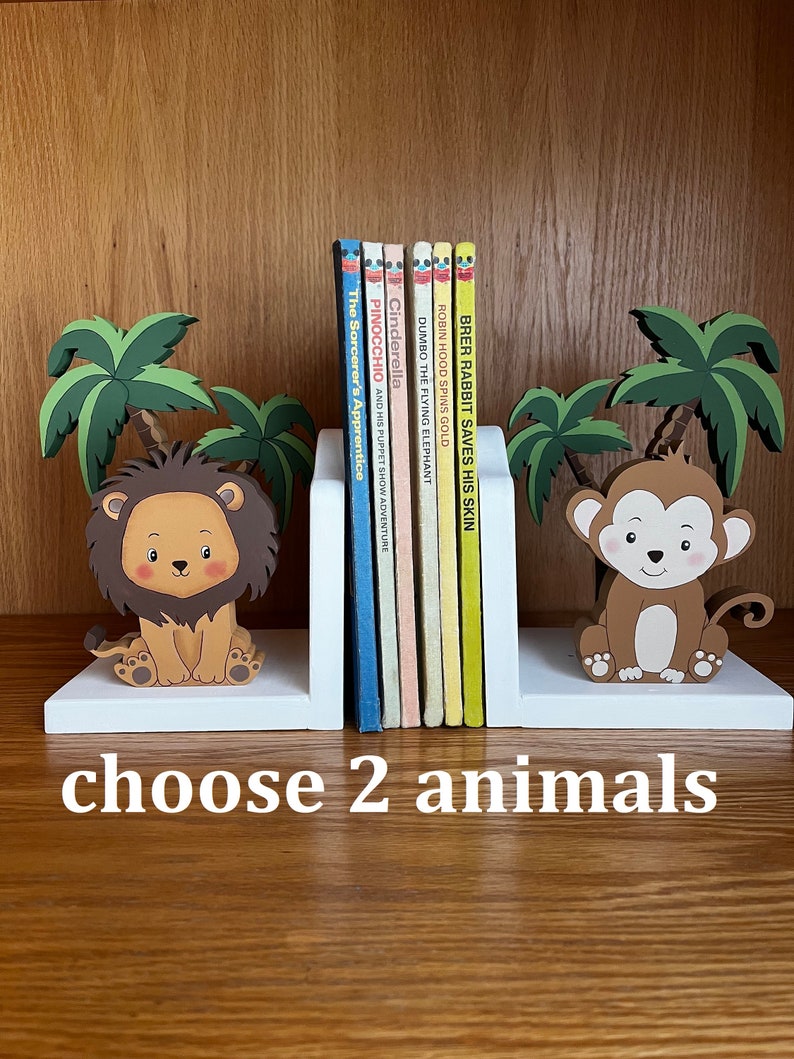 Peut inclure: Deux serre-livres en bois blanc avec une figurine de lion et de singe. Les serre-livres sont plac&eacute;s sur une &eacute;tag&egrave;re en bois avec une rang&eacute;e de livres pour enfants derri&egrave;re eux. Le texte sur les serre-livres indique "choisissez 2 animaux".