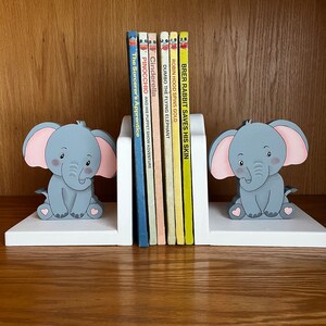 Peut inclure: Une paire de serre-livres en bois blanc en forme d'&eacute;l&eacute;phants avec des oreilles roses et des corps gris. Les serre-livres tiennent une rang&eacute;e de livres pour enfants color&eacute;s avec des titres comme "Brer Rabbit Saves His Skin", "Robin Hood Spins Gold", "Dumbo the Flying Elephant", "Cinderella", "Pinocchio", et "The Sorcerer's Apprentice".
