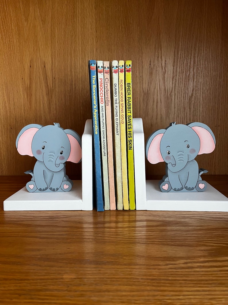 Peut inclure: Une paire de serre-livres en bois blanc en forme d'&eacute;l&eacute;phants avec des oreilles roses et des corps gris. Les serre-livres tiennent une rang&eacute;e de livres pour enfants color&eacute;s avec des titres comme "Brer Rabbit Saves His Skin", "Robin Hood Spins Gold", "Dumbo the Flying Elephant", "Cinderella", "Pinocchio", et "The Sorcerer's Apprentice".