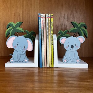 Peut inclure: Une paire de serre-livres en bois blanc repr&eacute;sentant un &eacute;l&eacute;phant gris et un koala gris. Les serre-livres sont d&eacute;cor&eacute;s de palmiers et de c&oelig;urs roses. Les serre-livres maintiennent une rang&eacute;e de livres pour enfants avec des titres tels que "Cendrillon", "Pinocchio", "Dumbo l'&eacute;l&eacute;phant volant", "Robin des Bois fait tourner l'or" et "Brer Lapin sauve sa peau".