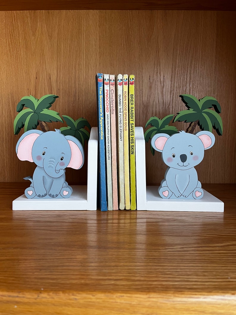 Peut inclure: Une paire de serre-livres en bois blanc repr&eacute;sentant un &eacute;l&eacute;phant gris et un koala gris. Les serre-livres sont d&eacute;cor&eacute;s de palmiers et de c&oelig;urs roses. Les serre-livres maintiennent une rang&eacute;e de livres pour enfants avec des titres tels que "Cendrillon", "Pinocchio", "Dumbo l'&eacute;l&eacute;phant volant", "Robin des Bois fait tourner l'or" et "Brer Lapin sauve sa peau".