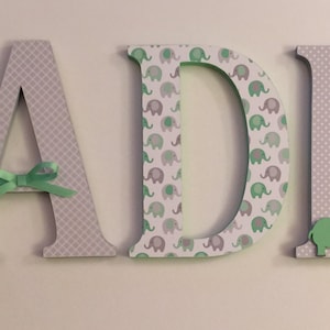 May include: Green and gray wooden letters spelling out the name "SADIE". The letters are decorated with a pattern of small elephants and a gray and white checkered pattern. A green bow is tied around the letter "A".