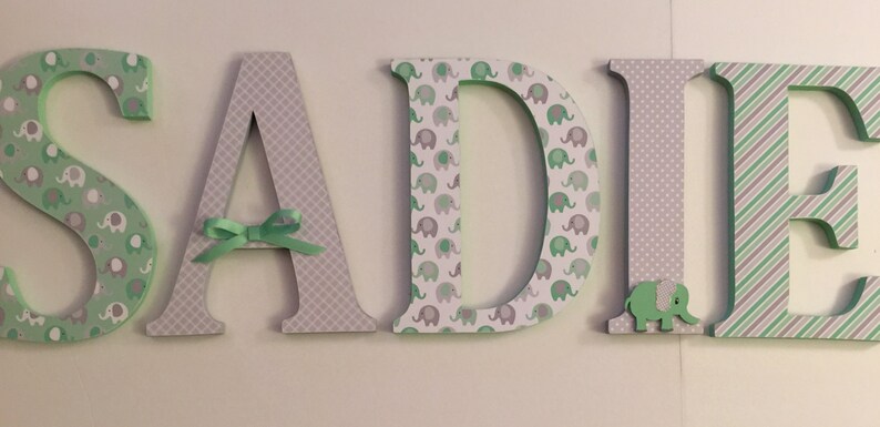 May include: Green and gray wooden letters spelling out the name "SADIE". The letters are decorated with a pattern of small elephants and a gray and white checkered pattern. A green bow is tied around the letter "A".