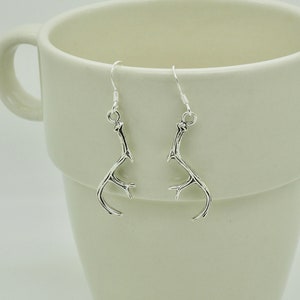 Op de afbeelding: Een paar zilverkleurige oorhangers in de vorm van een gewei. De oorbellen zijn ontworpen om op kleine herten geweien te lijken, met meerdere punten en een gebogen vorm. Ze hangen aan eenvoudige zilveren haken, wat een door de natuur geïnspireerd sieradenontwerp presenteert.