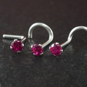 May include: Three silver nose studs with bright pink gemstones. Two studs have a curved post, and one has a straight post. The jewelry is displayed on a dark, textured surface.