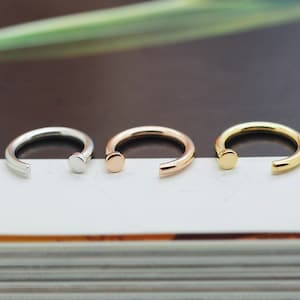 May include: Three open-hoop nose rings in silver, rose gold, and gold. Each ring has a small, flat, circular stud on one end. The rings are displayed on a white surface with a dark brown background.