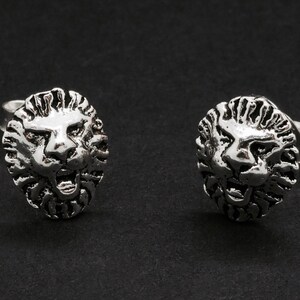 May include: Two silver-toned lion head stud earrings. Each earring features a detailed lion's face with a textured mane and open mouth. The earrings are set against a dark background.