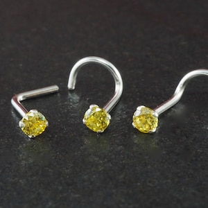 May include: Three silver nose studs, each featuring a bright yellow gemstone. The studs have a curved post design. The gemstones are round and set in a silver setting. The background is a dark, textured surface.