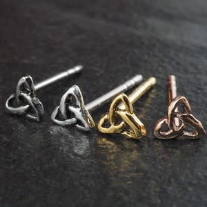 Puede incluir: Cuatro pendientes de botón con diseños de nudos celtas en tonos plata, oro y bronce. Los pendientes se exhiben sobre una superficie oscura y texturizada, resaltando los intrincados detalles de cada uno.