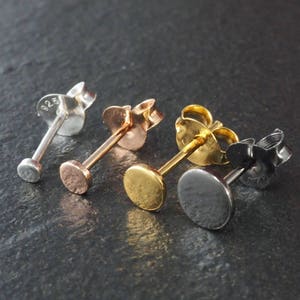 Op de afbeelding: Vier paar oorstekers in zilver, roségoud, goud en zwart. Elke oorbel heeft een kleine, platte, cirkelvormige stud en een vlindersluiting. De zilveren oorbel is gestempeld met "925".