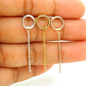 May include: Three delicate earrings in silver, gold, and rose gold tones. Each earring features a small circular hoop connected to a fine chain that dangles below. The earrings are displayed against a neutral background.