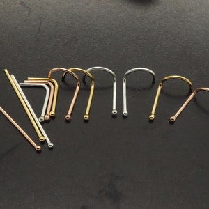 Op de afbeelding: Een assortiment metalen neusknopjes en neusringen in goud, zilver en roségoud tinten. De neusknopjes zijn recht met een kleine bal aan het uiteinde, terwijl de neusringen een gebogen ontwerp hebben. De sieraden worden op een donkere ondergrond getoond.