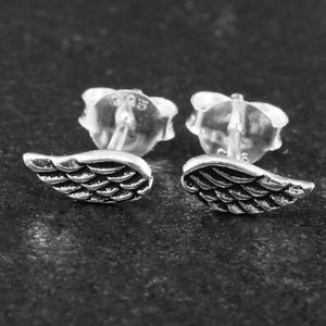 Op de afbeelding: Een paar zilverkleurige vleugeloorstekers. Elke oorbel heeft een gedetailleerd vleugelontwerp met getextureerde veerdetaillering. De oorbellen zijn geplaatst tegen een donkere, getextureerde achtergrond.