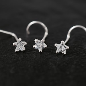 May include: Three silver nose studs, each featuring a star-shaped, clear cubic zirconia stone. The studs are displayed on a dark, textured surface. The star-shaped design adds a touch of sparkle and elegance to the jewelry.