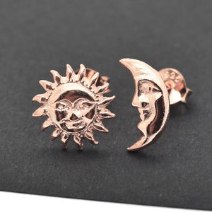 May include: Rose gold-colored stud earrings featuring a sun and a crescent moon design. The sun has a smiling face and radiating rays. The moon has a detailed face. The earrings are displayed on a dark surface.
