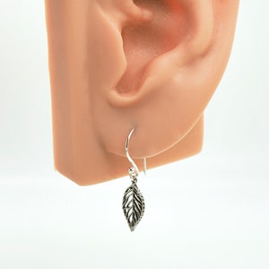 May include: A silver leaf earring with a hook, shown next to an ear. The leaf has detailed veining and a small, clear accent. The earring is hanging from a silver hook.