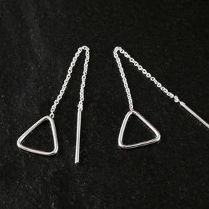 Puede incluir: Un par de pendientes colgantes plateados. Cada pendiente presenta un dije triangular suspendido de una delicada cadena, con una barra recta en el extremo. Se muestran sobre una superficie oscura y texturizada.