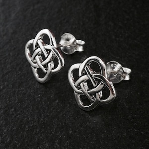 May include: A pair of silver-colored stud earrings. Each earring features a Celtic knot design, with interwoven lines forming a symmetrical pattern. The earrings are set against a dark, textured background.