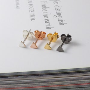 Può includere: Una collezione di quattro orecchini a bottone a triangolo in argento, oro rosa, oro e nero. Ogni orecchino presenta un piccolo design triangolare geometrico e un perno con chiusura a farfalla.