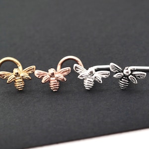 May include: Four bee-shaped nose studs in gold, rose gold, silver, and black. Each stud features a detailed bee design with wings and a curved or straight post. The studs are displayed on a dark surface.