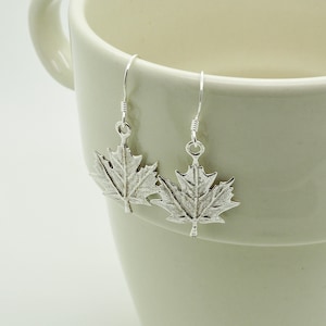 May include: A pair of silver maple leaf earrings. The earrings are suspended from a white mug. The earrings have a textured surface and are attached to simple hooks. The earrings are made of a silver-colored metal.