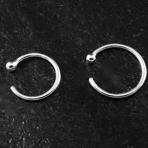 Può includere: Due anelli per il naso color argento, ciascuno con una piccola sfera a un'estremità e un design aperto. Gli anelli sono esposti su una superficie scura e strutturata, mostrando il loro design semplice ed elegante. Ideali per gioielli per il corpo.