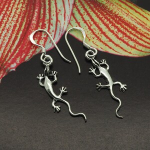 May include: A pair of silver lizard earrings. Each earring has a lizard pendant with detailed legs and a curled tail, hanging from a simple hook. The earrings are set against a dark background with a floral design.