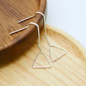 Puede incluir: Un par de pendientes de hilo plateados con un diseño de triángulo geométrico. Los pendientes presentan una delicada cadena y un pequeño colgante de triángulo abierto. Se muestran en una bandeja de madera.