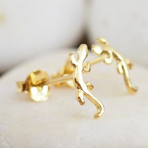 May include: A pair of gold-colored stud earrings. Each earring is shaped like a small lizard with detailed legs and tail. The earrings are displayed on a white surface.
