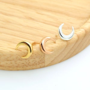 May include: Three crescent moon stud earrings in gold, rose gold, and silver. The earrings are displayed on a light wooden surface. The earrings are small and delicate, with a simple, elegant design. Jewelry.