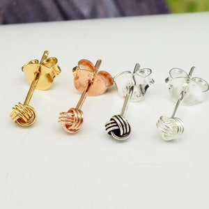 May include: Four pairs of stud earrings in gold, rose gold, black, and silver. Each earring features a knot design at the end of a post with a butterfly backing. The earrings are displayed against a light background.