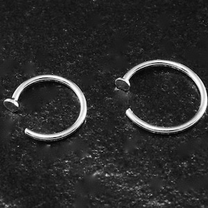Può includere: Due anelli per il naso argentati sono mostrati su uno sfondo scuro e strutturato. Ogni anello ha un design circolare con un piccolo disco piatto a un'estremità. Gli anelli sono aperti, progettati per essere inseriti in un piercing.