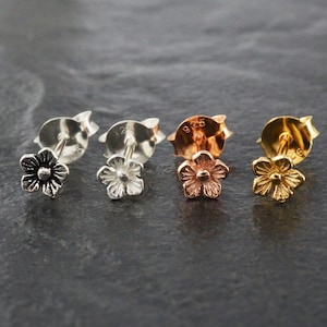 May include: Four pairs of flower-shaped stud earrings in silver, rose gold, and gold tones. The earrings are displayed on a dark grey surface. Each earring has a post and butterfly backing.