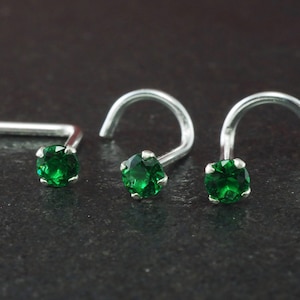 May include: Three silver nose studs, each featuring a round, emerald green gemstone. The studs are displayed with varying post designs, including curved and straight. The gemstones are set in a four-prong setting.