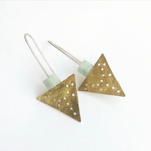 May include: A pair of brass triangle earrings with a textured finish and small circular cutouts. Each earring features a pale green square bead and a silver wire hook. The earrings are displayed on a white background.