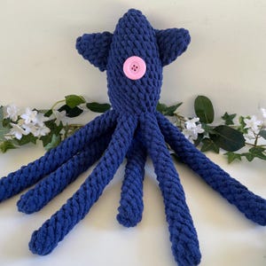 May include: A handmade, navy blue crocheted octopus plush toy. The plush features a pink button eye and eight long, textured tentacles. The octopus is displayed on a white surface with green and white floral accents.