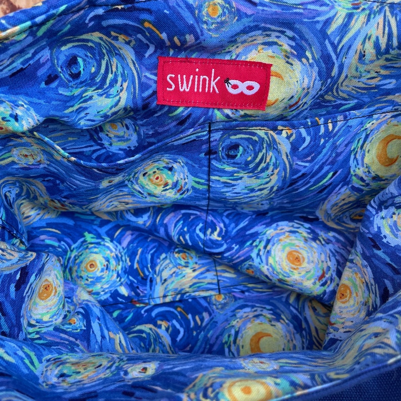 Puede incluir: El interior de una bolsa de lona azul con un patr&oacute;n de noche estrellada en espiral. La bolsa tiene una etiqueta roja con la palabra "swink" y un s&iacute;mbolo de infinito.