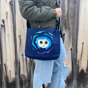 Puede incluir: Una bolsa de mensajero de lona azul con un gr&aacute;fico de la cara de una ni&ntilde;a con ojos grandes. La ni&ntilde;a tiene el pelo azul y lleva una flor rosa.