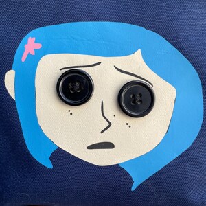 Puede incluir: Una ilustraci&oacute;n azul y blanca de una ni&ntilde;a con cabello azul y ojos negros en forma de botones. La ni&ntilde;a tiene una flor rosa en la cabeza y una expresi&oacute;n triste.
