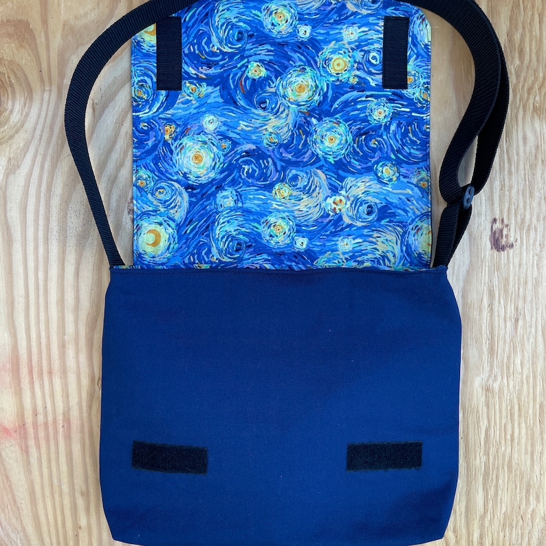 Puede incluir: Un bolso bandolera de lona azul con forro estampado de noche estrellada. El bolso tiene una correa ajustable negra y dos cierres de velcro negros.