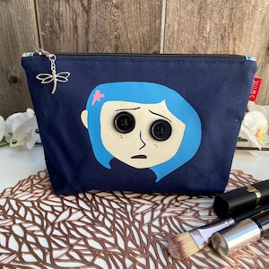 Puede incluir: Una bolsa de maquillaje de lona azul oscuro con una imagen impresa de una ni&ntilde;a con cabello azul y grandes botones negros para los ojos. La bolsa tiene una cremallera y un dije de lib&eacute;lula plateado.